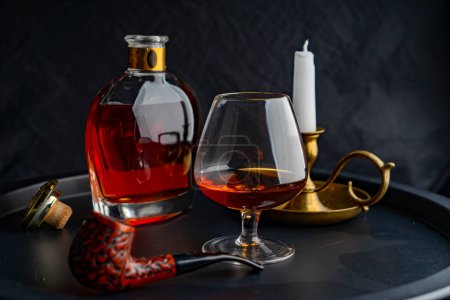 Téléchargez les photos : Un verre de cognac sur le fond d'une bouteille de cognac d'une bougie blanche dans un chandelier en bronze, sur un plateau sombre sur un fond sombre et une pipe pour fumer au premier plan - en image libre de droit