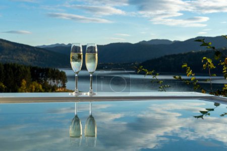 Téléchargez les photos : Deux verres de champagne au bord de la piscine sur fond de montagnes, lac, ciel bleu avec nuages - en image libre de droit
