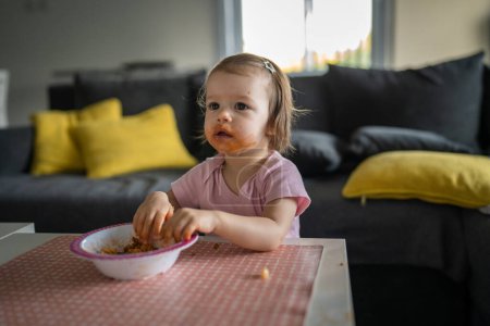 Téléchargez les photos : One girl small caucasian toddler female child daughter eating alone at the table at home while watching tv making mess on her face dirty sauce childhood growing up development concept copy space - en image libre de droit