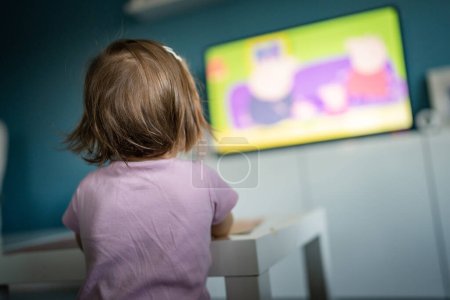 Back view of one unknown caucasian child toddler girl standing at home watching tv while eating copy space childhood growing up development leisure concept