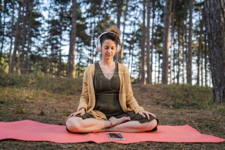 Una mujer joven adulta caucásica sentada sola en el parque o bosque en la naturaleza sosteniendo auriculares ajustables preparándose para la meditación guiada en línea auto-cuidado concepto de prácticas de equilibrio mental