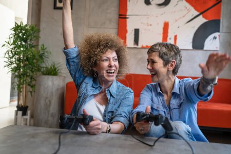Foto de Dos mujeres caucásicas amigas o hermanas jugando videojuegos en la consola en casa sosteniendo controladores de joystick divirtiéndose felices mujeres maduras personas reales copiar espacio celebrar la victoria - Imagen libre de derechos