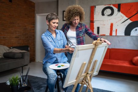 Téléchargez les photos : Deux femmes amies ou femme avec son mentor professeur d'art caucasien sœurs matures enseignent et apprennent à peindre aider pendant la peinture sur le chevalet à la maison dans un espace de copie de pièce lumineuse - en image libre de droit