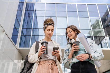 Photo for Two women female students walk in front of modern university building with notes and paper documents using mobile phone discuss talk happy smile while holding mobile phone reading sms text messages - Royalty Free Image