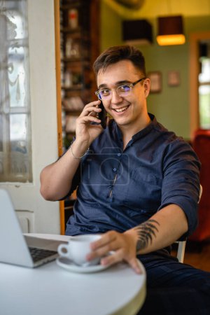 Téléchargez les photos : Un homme jeune homme caucasien assis dans le bureau à la maison ou au café travaillant sur ordinateur portable seul sourire projet freelance personnes réelles copier l'espace en utilisant téléphone mobile parler passer un appel - en image libre de droit