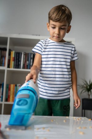 Foto de Un niño en edad preescolar limpieza desorden en la mesa con la aspiradora de mano después de jugar y hacer experimentos ayudando con la limpieza del hogar niño creciendo concepto - Imagen libre de derechos