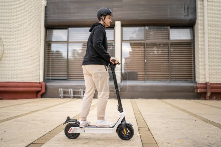 Foto de Un hombre joven adulto caucásico macho de pie en scooter de patada eléctrica en conducción de día en la ciudad o ciudad e-scooter ecológico modo de transporte personas reales copiar espacio longitud completa - Imagen libre de derechos