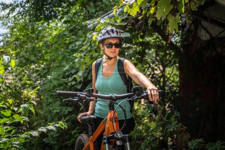 Foto de Una mujer caucásica adulta se prepara para montar bicicleta eléctrica en un día soleado de verano preparándose frente al arbusto verde, use casco protector y anteojos, la gente real copia el espacio - Imagen libre de derechos