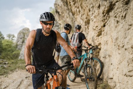Téléchargez les photos : Homme caucasien mâle cycliste équitation e-bike vélo électrique en plein air dans la chaîne de montagnes à la gorge ou la crête en face de ses amis portent un casque de protection et des lunettes dans l'espace de copie de jour ensoleillé - en image libre de droit