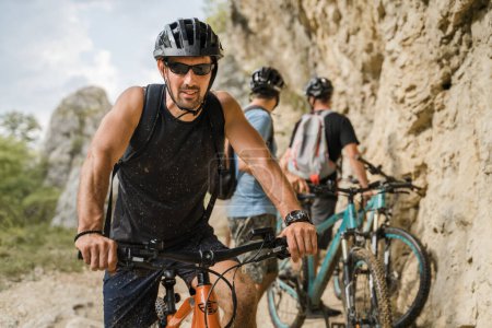 Téléchargez les photos : Homme caucasien mâle cycliste équitation e-bike vélo électrique en plein air dans la chaîne de montagnes à la gorge ou la crête en face de ses amis portent un casque de protection et des lunettes dans l'espace de copie de jour ensoleillé - en image libre de droit