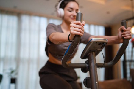 Photo for One woman young caucasian female training at home with headphones on Indoor Cycling stationary Exercise Bike real people copy space health and fitness workout concept selective focus on hand - Royalty Free Image