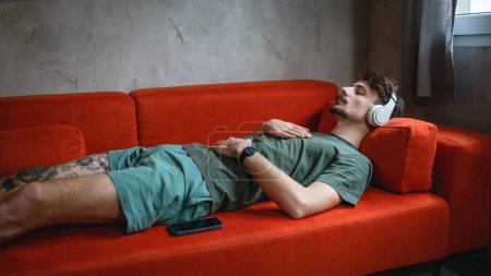 Téléchargez les photos : Un homme adulte caucasien mâle sur le canapé-lit utilisent des écouteurs pour la méditation guidée en ligne pratiquant le yoga pleine conscience avec les yeux fermés à la maison de vraies personnes self care manifestation concept copier l'espace - en image libre de droit