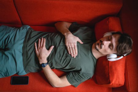 Téléchargez les photos : Un homme adulte caucasien mâle sur le canapé-lit utilisent des écouteurs pour la méditation guidée en ligne pratiquant le yoga pleine conscience avec les yeux fermés à la maison de vraies personnes self care manifestation concept copier l'espace - en image libre de droit