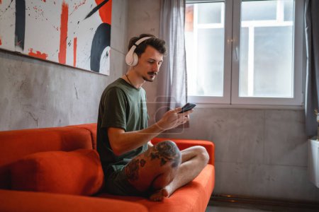 Téléchargez les photos : One man sit on the bed at home with headphones use smart-phone for music listening or search for podcast online meditation or audiobook on smarphone mobile phone online on internet copy space - en image libre de droit