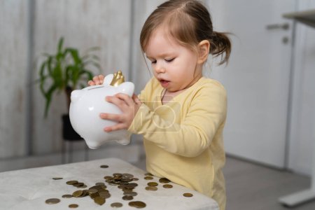 Foto de Una pequeña chica caucásica de dos años de edad jugar con monedas y alcancía en casa seguro de ahorro de dinero y el futuro concepto de inversión - Imagen libre de derechos