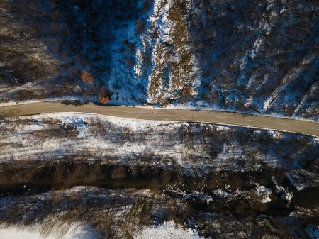 Téléchargez les photos : Vue aérienne sur drone sur la route à travers la colline et la montagne dans la journée d'hiver couverte de neige avec roche et arbres et rivière - en image libre de droit