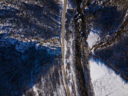 Téléchargez les photos : Vue aérienne sur drone sur la route à travers la colline et la montagne dans la journée d'hiver couverte de neige avec roche et arbres et rivière - en image libre de droit