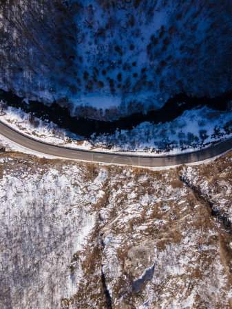 Téléchargez les photos : Vue aérienne sur drone sur la route à travers la colline et la montagne en journée d'hiver couverte de neige avec des rochers et des arbres et rivière en Serbie - en image libre de droit