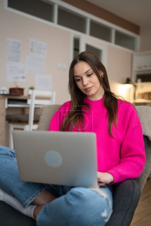 Téléchargez les photos : Une jeune femme utilise un ordinateur portable à la maison tout en étant assise confortablement - en image libre de droit