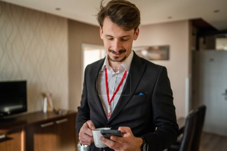 Foto de One man adult caucasian businessman wear suit in hotel room while taking a brake or prepare for work hold cup of coffee and smartphone sending sms messages copy space - Imagen libre de derechos