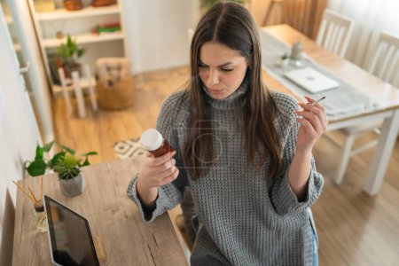 Foto de One woman young caucasian female hold medicine drugs pills tablets while siting at home smoke cigarette thinking trying to quit smoking real person copy space - Imagen libre de derechos