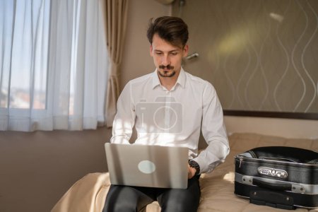 Foto de One man adult caucasian businessman sit on the bed in hotel room work on his laptop computer prepare for meeting or remote online conference real person copy space wear white shirt - Imagen libre de derechos