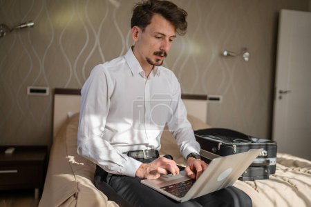 Téléchargez les photos : Un homme adulte caucasien homme d'affaires assis sur le lit dans la chambre d'hôtel travailler sur son ordinateur portable se préparer pour réunion ou conférence en ligne à distance espace de copie personne réelle porter chemise blanche - en image libre de droit