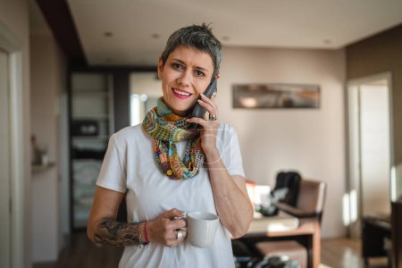 Photo for One woman mature senior caucasian female with modern short gray hair stand at home in room use mobile smart phone to make a call talking daily morning routine real people copy space hold cup of coffee - Royalty Free Image