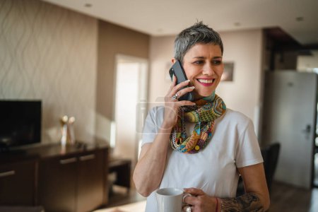Foto de One woman mature senior caucasian female with modern short gray hair stand at home in room use mobile smart phone to make a call talking daily morning routine real people copy space hold cup of coffee - Imagen libre de derechos