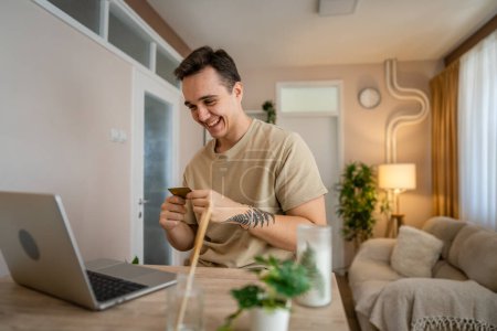 One man adult caucasian male sit at home happy smile at laptop computer hold bank credit card online shopping concept real people copy space e-commerce