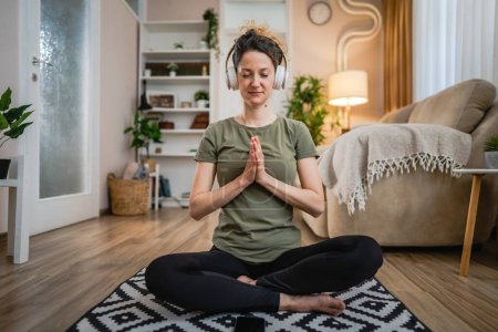 Téléchargez les photos : Une femme adulte caucasien femelle millénium en utilisant des écouteurs pour la méditation guidée en ligne pratiquant le yoga pleine conscience avec les yeux fermés assis sur le sol à la maison personnes réelles espace de copie concept d'auto-soins - en image libre de droit