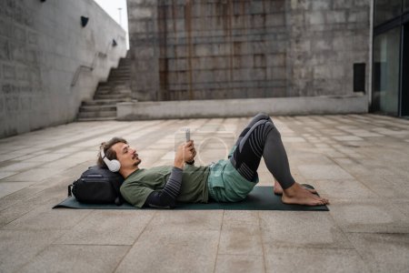 Téléchargez les photos : Un homme jeune adulte caucasien yeux masculins fermés pour la formation guidée yoga ou méditation tout en étant assis en plein air avec casque pratique d'auto-soins de vraies personnes bien-être concept de paix intérieure et d'équilibre - en image libre de droit