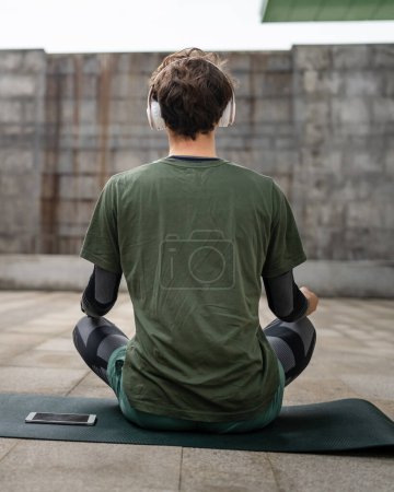 Un hombre joven adulto caucásico ojos masculinos cerrados para entrenamiento guiado yoga o meditación mientras está sentado al aire libre con auriculares auto-cuidado práctica personas reales bienestar paz interior y equilibrio concepto