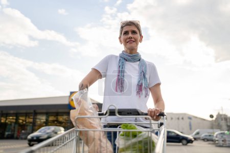 Foto de One mature woman caucasian female walking in the parking lot in front of the shopping center grocery store supermarket pushing chart happy smile half length copy space - Imagen libre de derechos