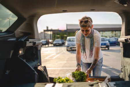 Foto de Una mujer madura caucásica hembra de pie junto al maletero trasero de su coche en el estacionamiento del supermercado centro comercial o tienda de comestibles con verduras comida en caja ponerlos en el vehículo - Imagen libre de derechos