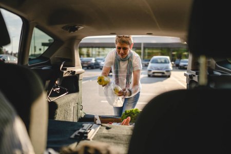 Foto de Una mujer madura caucásica hembra poner caja con verduras en la parte posterior de su coche en el día soleado en el estacionamiento frente al supermercado o tienda de comestibles en el espacio de copia día soleado personas reales - Imagen libre de derechos