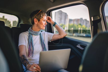 Photo for One mature woman caucasian female sitting on the back seat of the car thinking while looking to the side and working on laptop computer in summer day with short gray hair modern manager copy space - Royalty Free Image