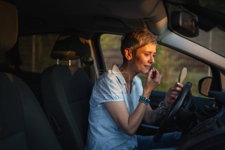 Photo for One woman mature caucasian female businesswoman sitting in car putting lipstick fixing repairing makeup on her face while waiting in summer day evening real people copy space gray hair - Royalty Free Image