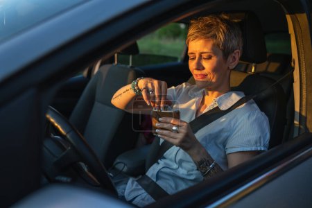 Téléchargez les photos : One woman mature caucasian female with tattoo on arms hold flask while sitting in the car alone driver on parked automobile drinking alcohol abuse and alcoholism concept copy space side view - en image libre de droit