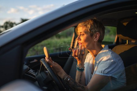 Téléchargez les photos : One woman mature caucasian female businesswoman sitting in car putting lipstick fixing repairing makeup on her face while waiting in summer day evening real people copy space gray hair - en image libre de droit