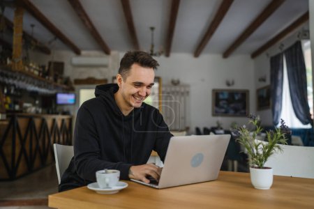 Photo for One man caucasian adult male sitting at the table at cafe restaurant wear black hoodie using computer laptop to browse internet or work freelance remote real people copy space happy smile successful - Royalty Free Image
