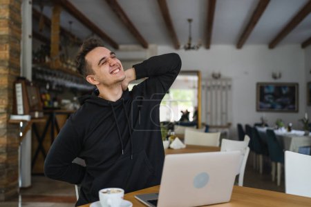 Photo for One man caucasian adult male sitting at the table at cafe restaurant wear black hoodie using computer laptop to browse internet or work real people copy space hold neck having pain back or neck strain - Royalty Free Image