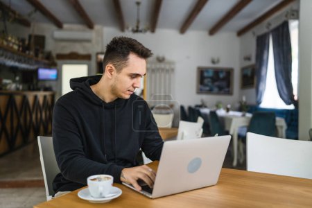 Foto de One man caucasian adult male sitting at the table at cafe restaurant wear black hoodie using computer laptop to browse internet or work freelance remote real people copy space - Imagen libre de derechos