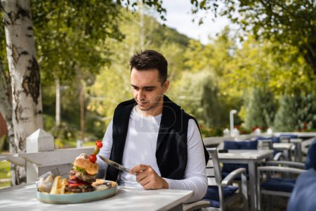 Téléchargez les photos : Un homme jeune homme caucasien adulte assis à la table au restaurant terrasse extérieure avec burgher et des croustilles de pommes de terre feu français manger seul dans la journée les gens réels copier l'espace - en image libre de droit