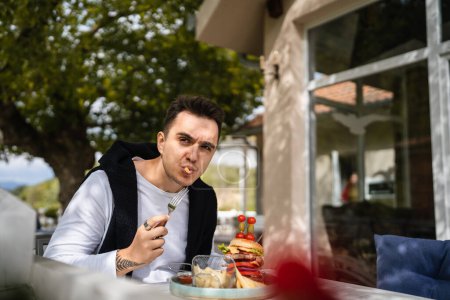 Téléchargez les photos : Un homme jeune homme caucasien adulte assis à la table au restaurant terrasse extérieure avec burgher et des croustilles de pommes de terre feu français manger seul dans la journée les gens réels copier l'espace - en image libre de droit