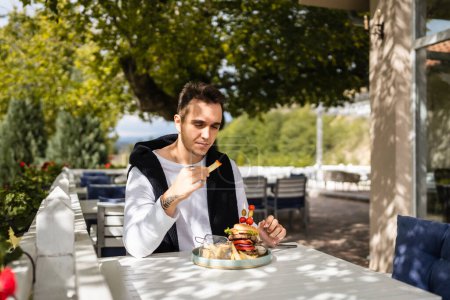 Photo for One man young adult caucasian male sitting at the table at restaurant outdoor terrace with burgher and french fires potato chips eat alone in day real people copy space - Royalty Free Image