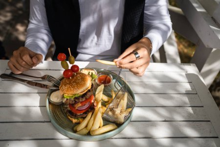 Téléchargez les photos : Gros plan sur le hamburger dans une assiette et les mains d'un homme caucasien inconnu se préparent à manger sur la table par une journée ensoleillée en plein air au restaurant - en image libre de droit