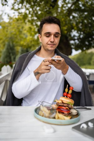 Photo for One man caucasian male sitting at terrace of the restaurant using mobile phone to take photo of food burger in a plate on the table in day outdoor real people copy space - Royalty Free Image