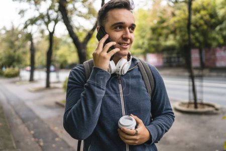 Photo for One young adult caucasian man walking in the city town near park using mobile phone smartphone talk with cup of coffee in autumn or spring day happy male tourist standing alone real people copy space - Royalty Free Image