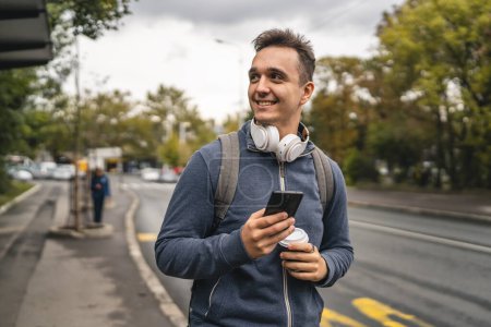 Photo for One man young adult caucasian male standing outdoor in the city waiting for the public bus transport at the station modern tourist or student in autumn day hold mobile phone real people copy space - Royalty Free Image